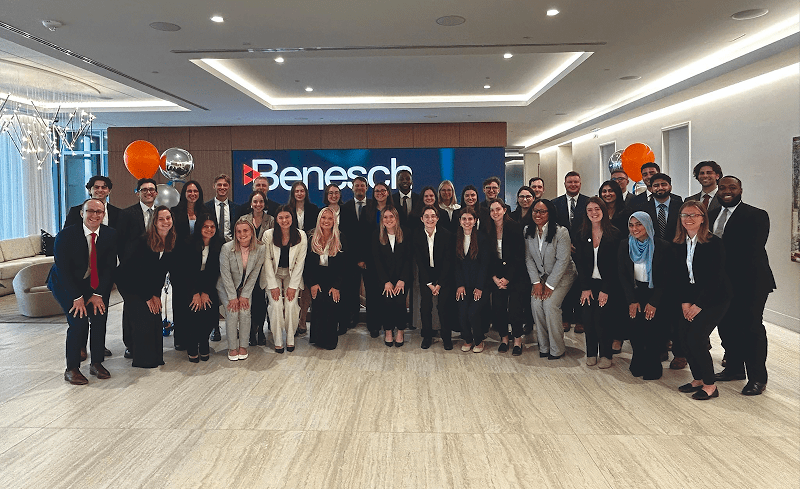 A large, diverse group of approximately 30 smiling people, mostly young professionals wearing business attire, poses for a group photo indoors. They are standing in front of a blue backdrop featuring the Benesch firm logo, with two metallic orange and silver balloon bouquets visible on either side of the display.