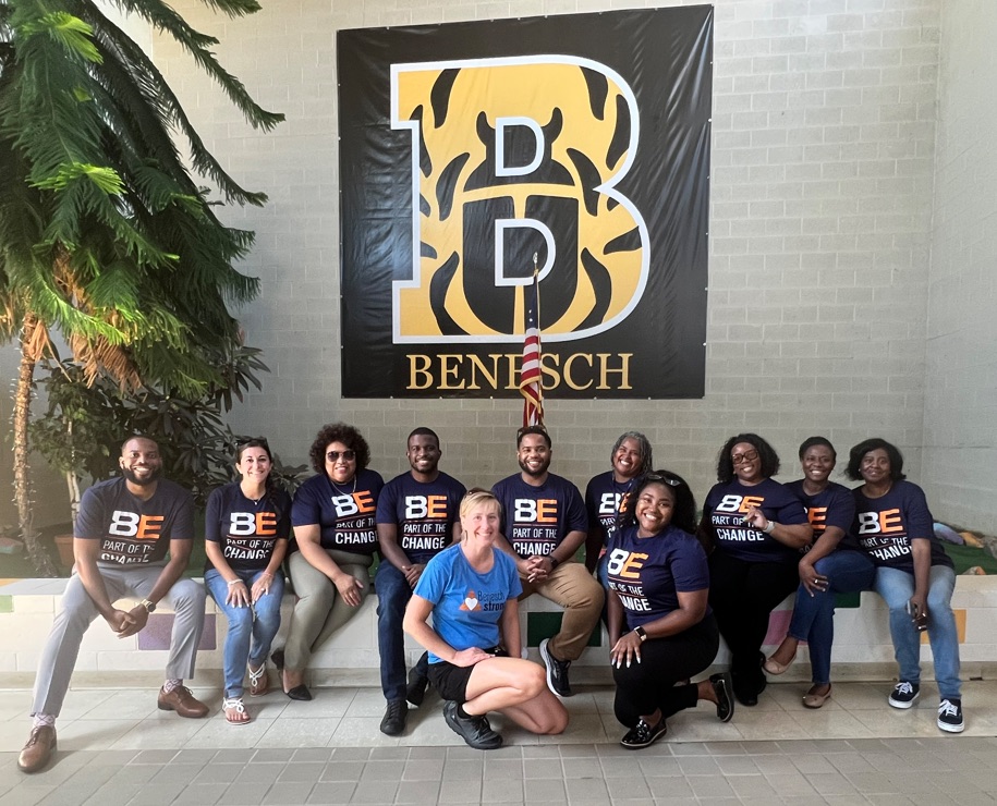 A group of eleven diverse people posing in front of a large black banner featuring a yellow "B" logo and the word "BENESCH". Most are wearing navy blue t-shirts that say "BE PART OF THE CHANGE," while one person in a blue shirt kneels in the front center.