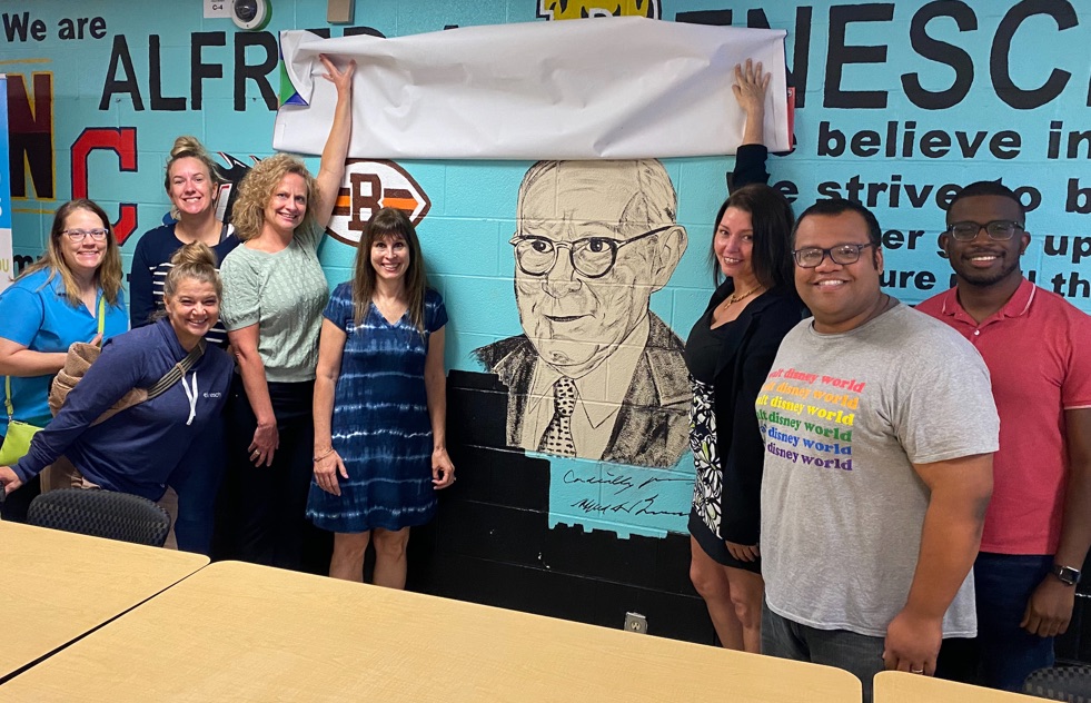 A group of eight people posing in a room next to a wall mural of a man's face. Two people hold up a white banner to reveal the top of the mural. The wall features the text "Alfred Benesch" and other motivational phrases.