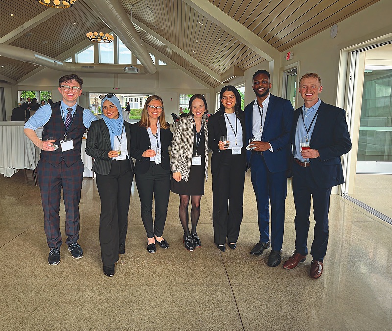 Seven smiling young professionals stand together in a bright, modern event space with high vaulted ceilings and polished floors. The group is dressed in business attire—including suits, blazers, and a blue hijab—and several individuals are holding drinks and wearing event lanyards.