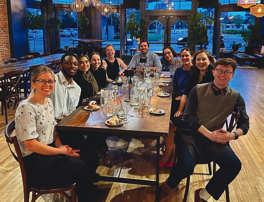 A group of ten diverse young professionals sitting around a long wooden table in a dimly lit, modern restaurant with large windows and brick walls. They are smiling and looking toward the camera, with half-eaten desserts and drinks on the table in front of them.